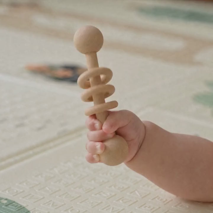 Main de bébé qui secoue le hochet en bois à trois anneaux au-dessus d'un tapis de jeu ECLOA